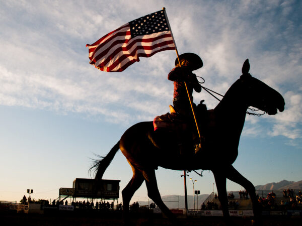 Silhouette of a person on horseback holding an American flag against a dusk sky in a rodeo arena. visit bishop