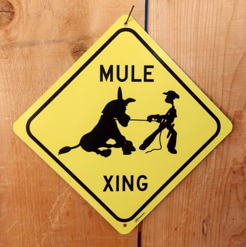 A diamond-shaped yellow sign with the text "MULE XING" evokes the spirit of Bishop, California. It features a black silhouette of a cowboy leading a mule with a rope and is attached to a wooden surface with a hook. visit bishop