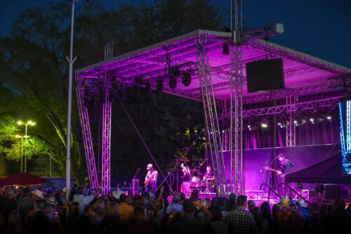 Nighttime outdoor concert with a band performing on a brightly lit stage and a crowd of people watching. visit bishop