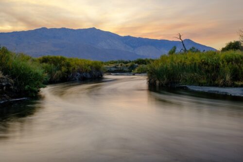 A tranquil river meanders through grassy banks near Bishop, California, with the majestic Eastern Sierra mountains and a softly lit sky in the background. visit bishop