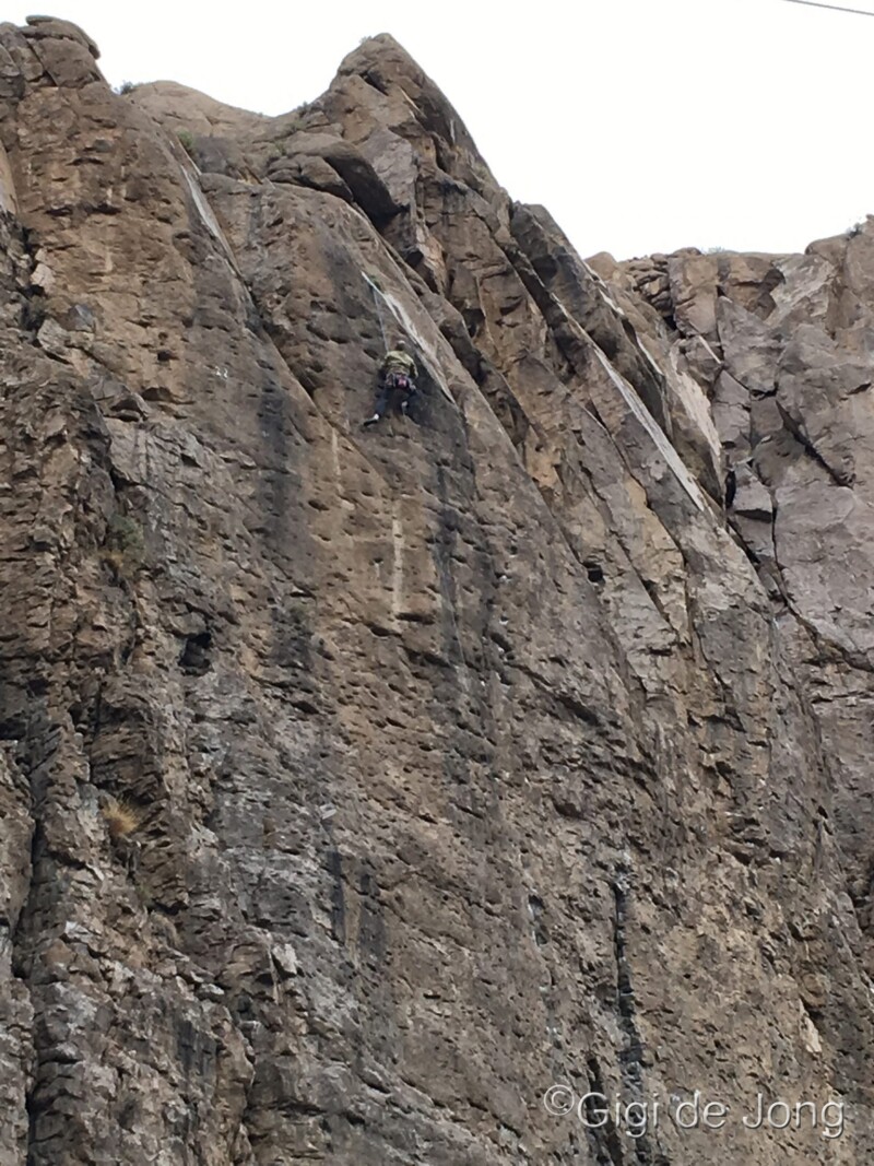 A rock climber scales a steep, rocky cliff face. The text "© Gigi de Jong" is in the bottom right corner. visit bishop