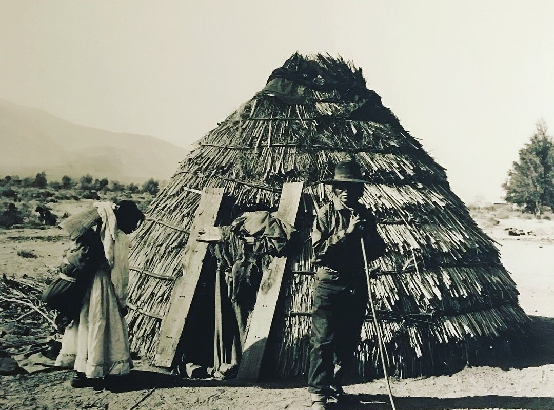 A vintage black-and-white photo from Bishop, California, depicts two individuals standing outside a round, thatched-roof hut made of straw and brush. One person appears to be leaning on a cane, while the other is partially bent over, with clothing and items hanging on a wooden structure nearby. visit bishop