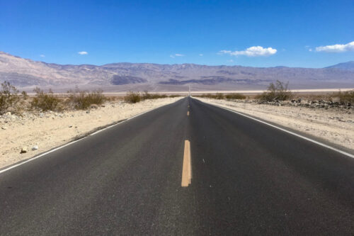 A straight, empty road stretches through a desert landscape with mountains in the distance under a clear blue sky. visit bishop