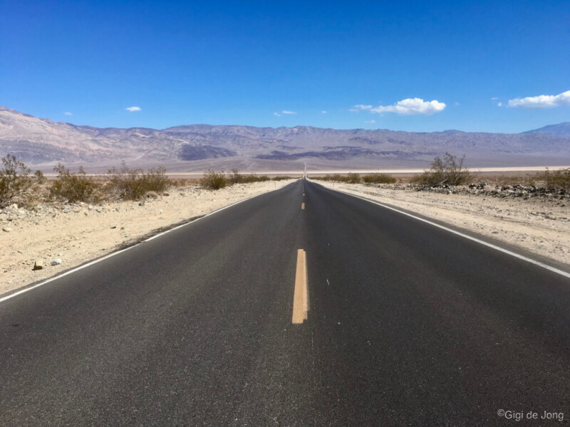 A straight, empty road stretches through a desert landscape with mountains in the distance under a clear blue sky. visit bishop