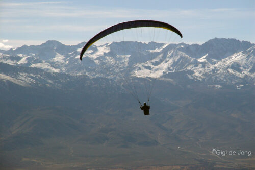 Person paragliding with mountainous landscape in the background. The sky is clear, and snow-covered peaks are visible. visit bishop