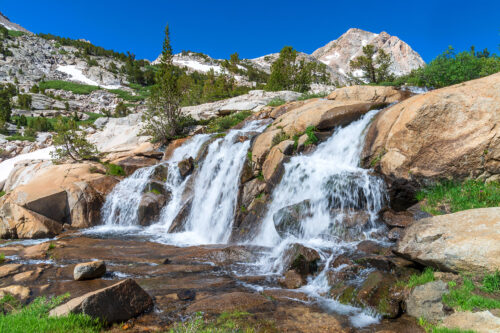 A cascading waterfall flows over rocks surrounded by greenery and mountains under a clear blue sky. visit bishop