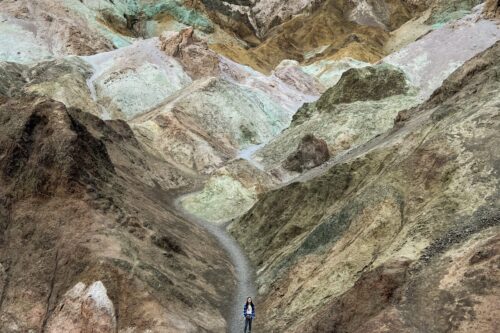 A person walks a narrow path in a colorful, rocky landscape under a blue sky. visit bishop
