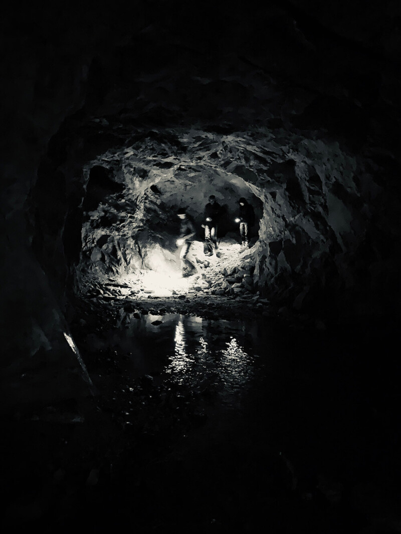 Three people exploring a dark, rocky cave with a shallow water stream on the ground, illuminated by headlamps. visit bishop