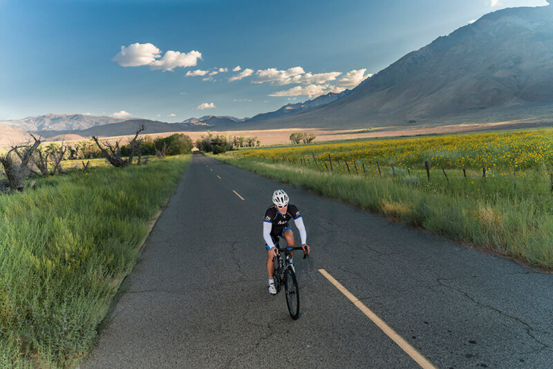 A cyclist wearing a helmet and cycling gear rides on an empty road near Bishop, California, surrounded by fields of green grass and yellow flowers. Mountains loom in the background under a partly cloudy sky. visit bishop