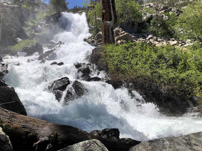 A powerful waterfall cascades over rocky terrain surrounded by lush greenery under a clear blue sky, evoking the breathtaking landscapes of Bishop, California. visit bishop