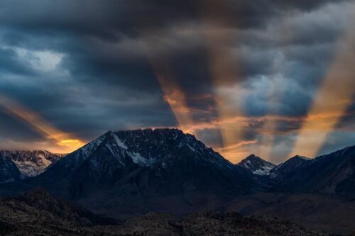 Sun rays breaking through dark clouds over a range of snowy, rugged mountains during sunset. visit bishop