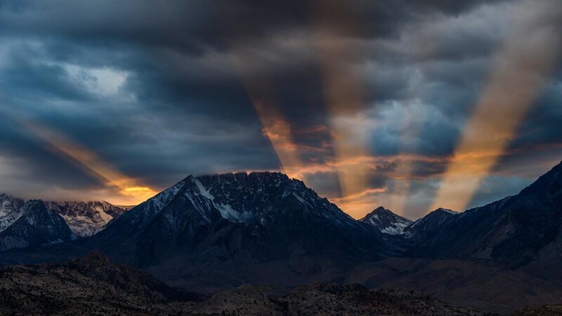 Sun rays breaking through dark clouds over a range of snowy, rugged mountains during sunset. visit bishop