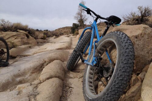 A blue fat-tire bike is parked on a rocky, uneven trail with a dry, barren landscape in the background. visit bishop