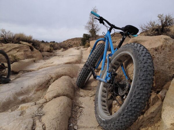 A blue fat-tire bike is parked on a rocky, uneven trail with a dry, barren landscape in the background. visit bishop