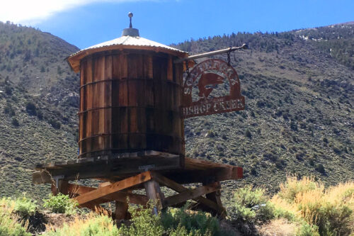 Old wooden water tank on elevated supports with an antique sign against a backdrop of hilly landscape under a blue sky. visit bishop