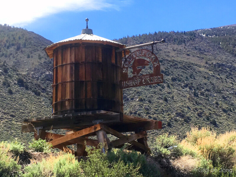 Old wooden water tank on elevated supports with an antique sign against a backdrop of hilly landscape under a blue sky. visit bishop