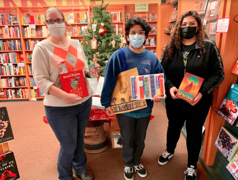 Three people are standing in a bookstore in Bishop, California, holding books. Two of them are wearing masks and glasses, and one is holding an object with multiple pens attached. The background includes a decorated Christmas tree and shelves filled with books. visit bishop