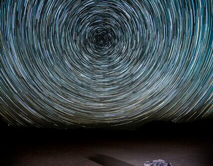 A dramatic time-lapse photograph shows star trails circling the night sky above a flat, barren desert landscape near Bishop, California. A solitary rock sits on the ground, casting a long shadow. The swirling star patterns create a hypnotic effect against the dark sky. visit bishop