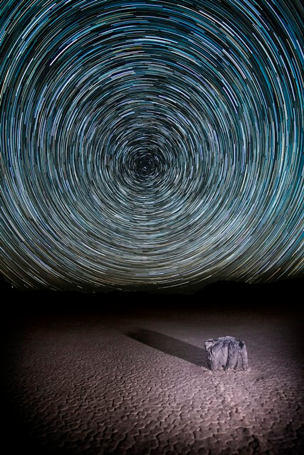 A dramatic time-lapse photograph shows star trails circling the night sky above a flat, barren desert landscape near Bishop, California. A solitary rock sits on the ground, casting a long shadow. The swirling star patterns create a hypnotic effect against the dark sky. visit bishop