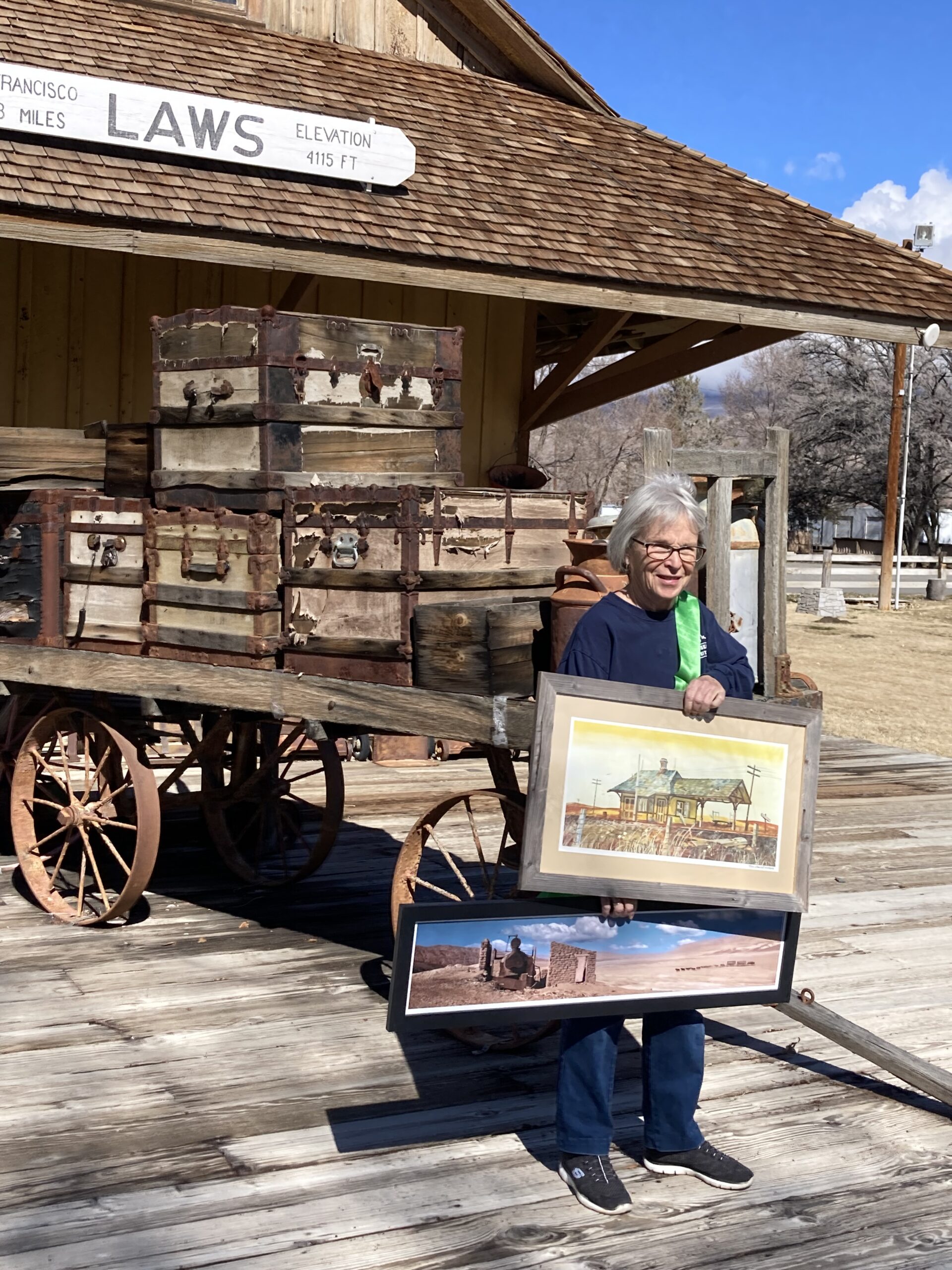 An elderly person holds framed photos next to an old wooden wagon stacked with vintage trunks, near a building labeled "LAWS" in Bishop, California, nestled in the heart of the Eastern Sierra. visit bishop