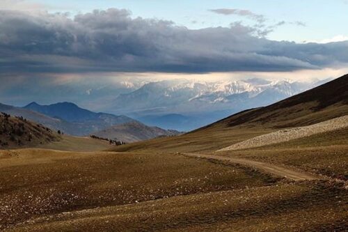 Rolling hills with a winding dirt path under a cloudy sky. Snow-capped mountains are visible in the distance. visit bishop