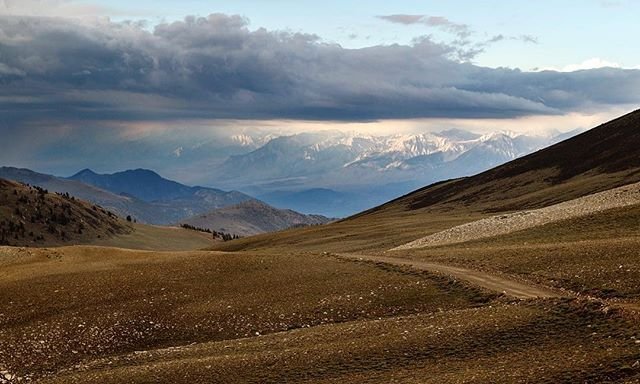 Rolling hills with a winding dirt path under a cloudy sky. Snow-capped mountains are visible in the distance. visit bishop