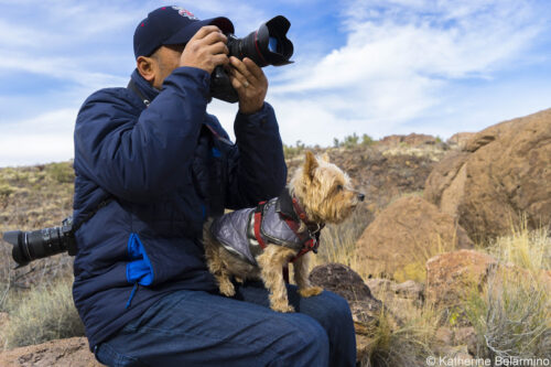 A person in a blue jacket and cap sits on a rocky outcrop in Bishop, California, holding a camera up to their face. A small dog in a gray jacket sits on their lap, both looking intently at something in the distance. Dry grass and rocky terrain are visible in the background. © Katherine Belarmino. visit bishop