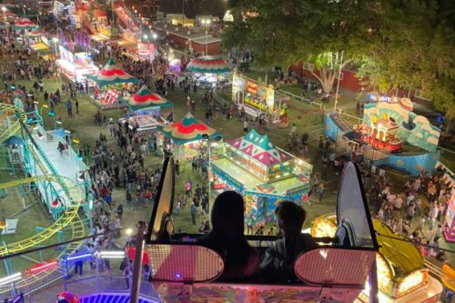 A nighttime aerial view of a busy carnival in Bishop, California, featuring brightly lit rides, game stalls, and a Ferris wheel in the background. In the foreground, two people are sitting in a ride, overlooking the lively scene below. visit bishop