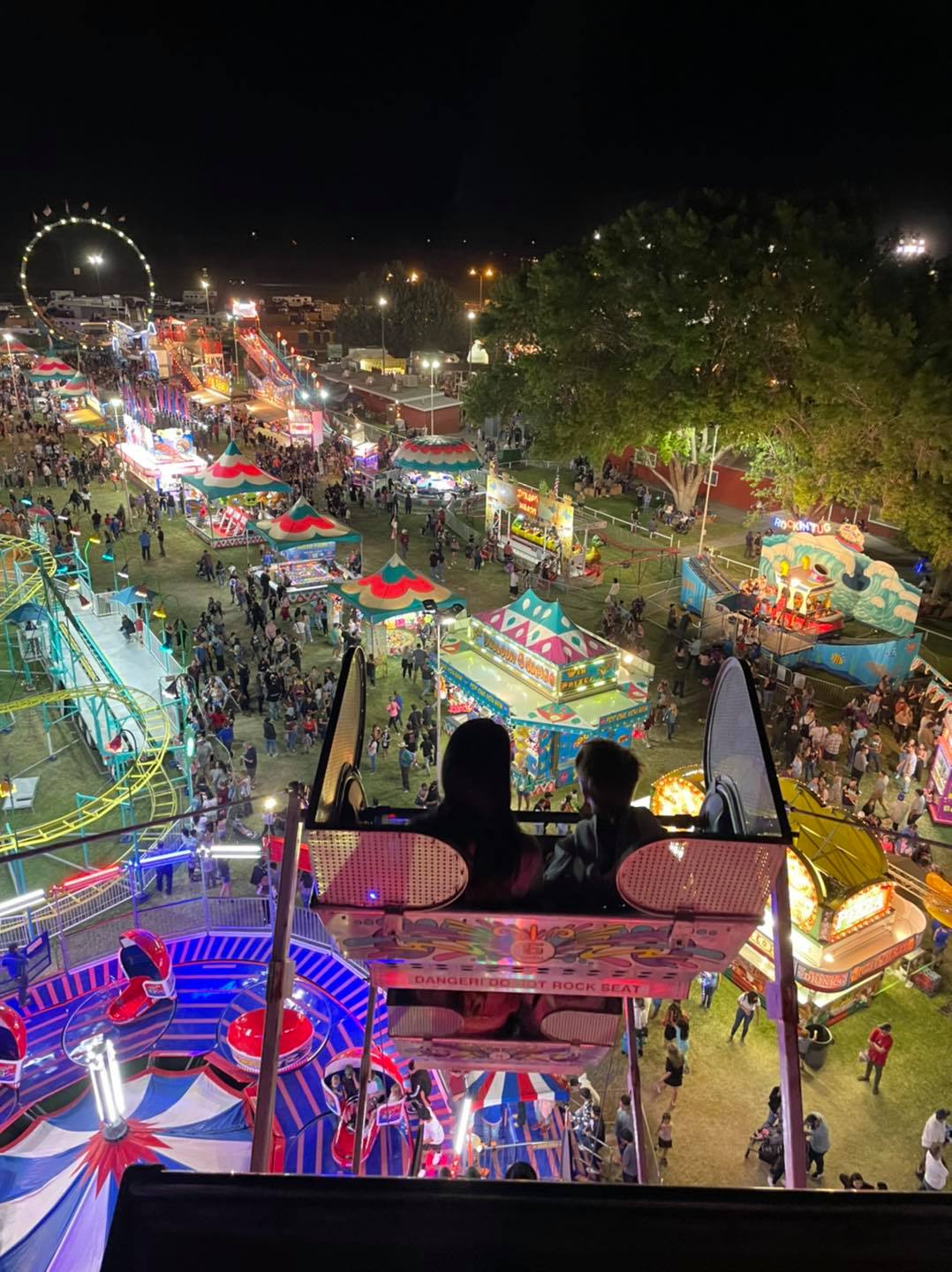 A nighttime aerial view of a busy carnival in Bishop, California, featuring brightly lit rides, game stalls, and a Ferris wheel in the background. In the foreground, two people are sitting in a ride, overlooking the lively scene below. visit bishop