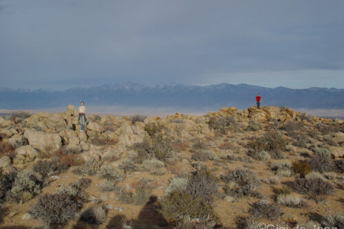 Two people stand on rocky desert terrain with distant mountains under a cloudy sky. Photo credit: Gigi de Jong. visit bishop