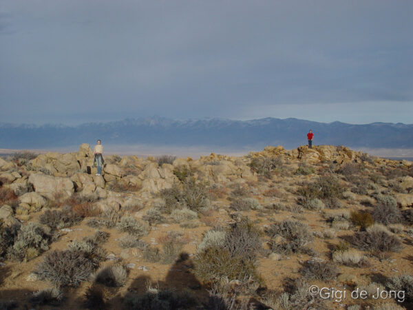 Two people stand on rocky desert terrain with distant mountains under a cloudy sky. Photo credit: Gigi de Jong. visit bishop