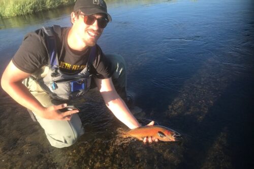 A person wearing a cap and fishing gear kneels in a river, holding a fish with a smile on their face. visit bishop