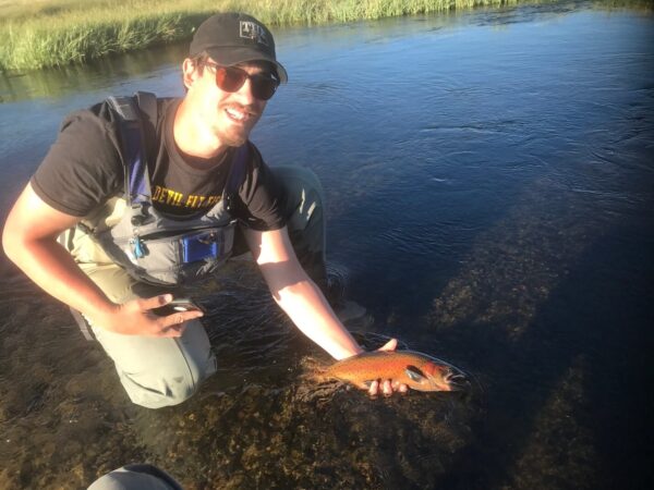 A person wearing a cap and fishing gear kneels in a river, holding a fish with a smile on their face. visit bishop