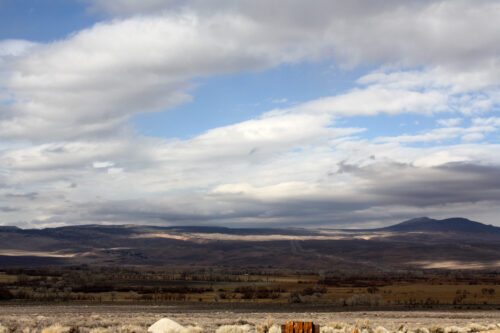 Expansive landscape with rolling hills, patches of sunlight, cloudy sky, and distant mountains on the horizon. visit bishop