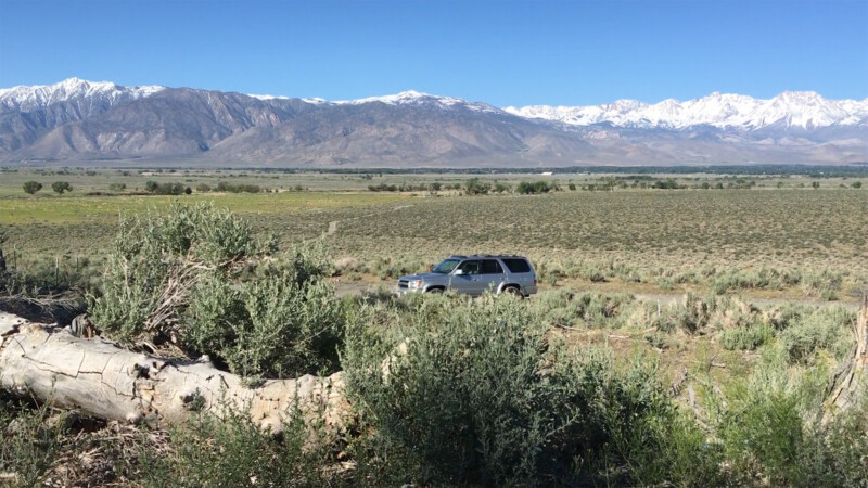 A silver SUV drives across a vast, arid landscape with snow-capped mountains in the background. visit bishop