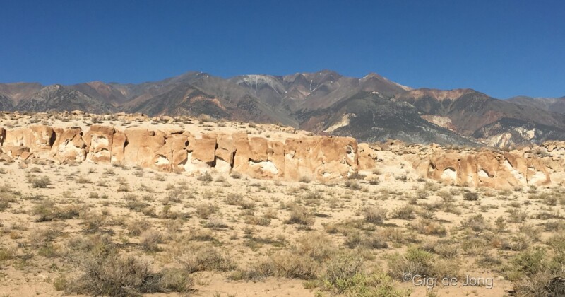 Rock formations and sparse vegetation in a desert landscape with mountains under a clear blue sky in the background. visit bishop