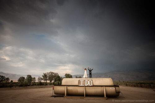 A newlywed couple stands on top of a large cylindrical water tank in Bishop, California, with "I DO" painted on it in bold white letters. The couple has their arms raised victoriously. The background shows a moody sky with mountains and trees in the distance. visit bishop