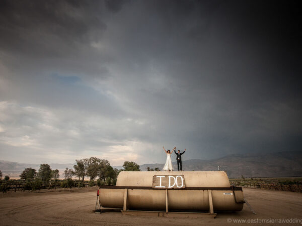 A newlywed couple stands on top of a large cylindrical water tank in Bishop, California, with "I DO" painted on it in bold white letters. The couple has their arms raised victoriously. The background shows a moody sky with mountains and trees in the distance. visit bishop