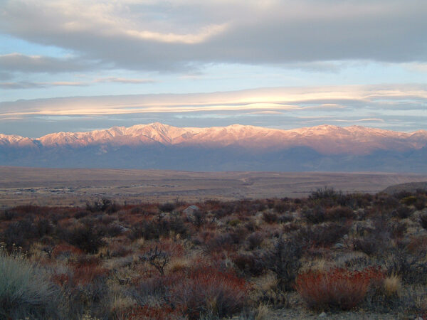 Mountain range with snow-covered peaks under a cloudy sky, viewed over a dry, shrub-filled landscape. visit bishop