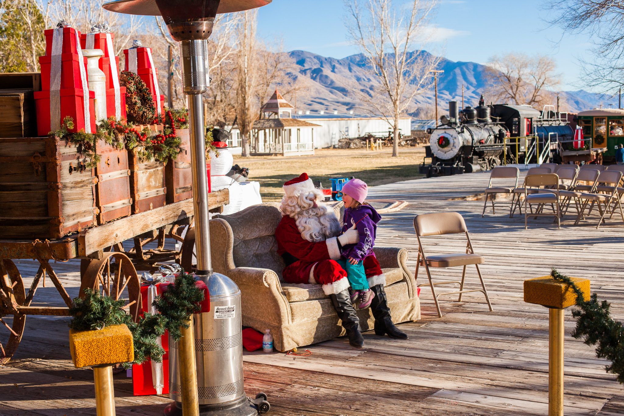 Santa Claus sitting on a bench talking with a child in a festive outdoor setting with the majestic Eastern Sierra mountains in the background. visit bishop