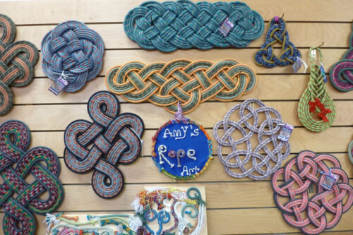 Various rope art pieces in different shapes and colors displayed on a wooden wall with a sign that reads "Amy's Rope Art. visit bishop