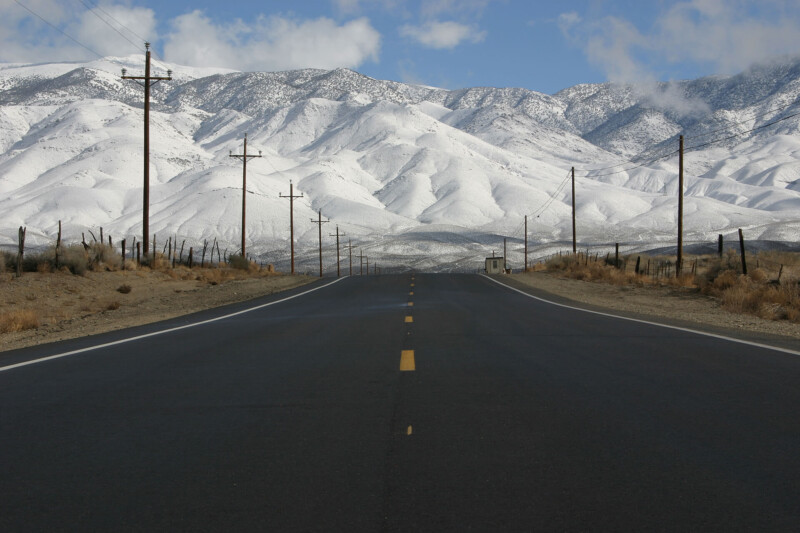 Empty road leading towards snow-covered mountains under a cloudy sky, with utility poles and fences on each side. visit bishop