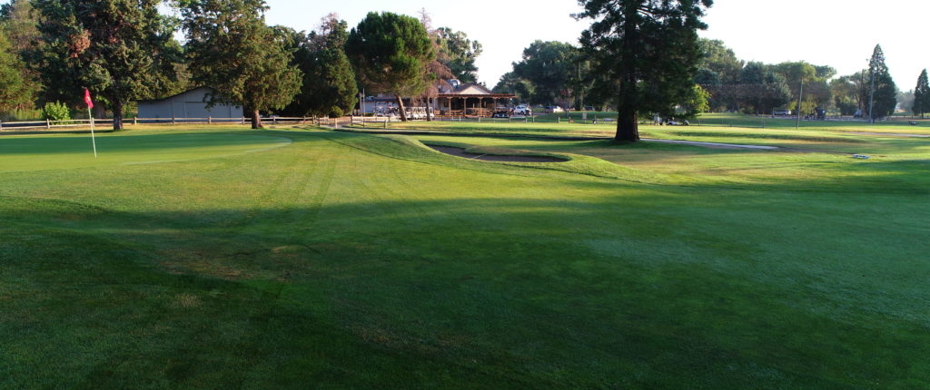 A serene view of a lush green golf course with a flagstick, trees, and a clubhouse in the background, nestled in the picturesque setting of Bishop, California, against the stunning Eastern Sierra backdrop. visit bishop