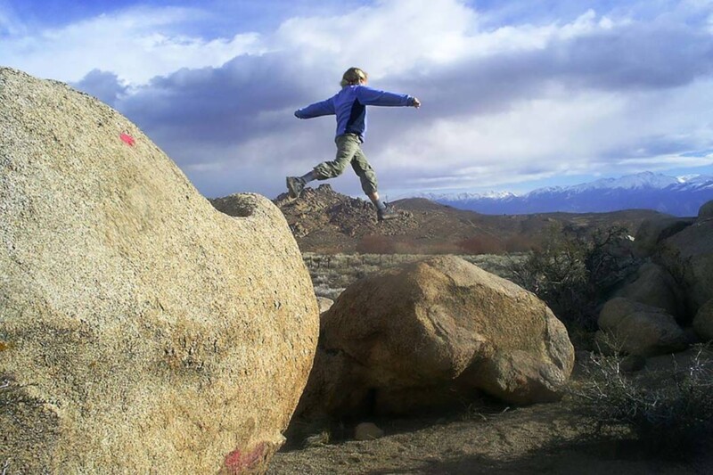 A person in a blue jacket and khaki pants is caught mid-air as they jump from one large rock to another in the rocky, desert landscape of Bishop, California. Snow-capped mountains and cloudy skies are visible in the background. visit bishop