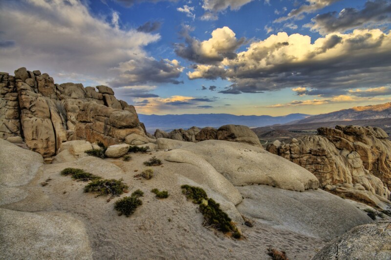 A rugged landscape of large, sunlit rock formations under a dramatic sky with clouds. Sparse vegetation dots the rocky terrain of Bishop, California, and distant mountains stretch across the horizon, illuminated by the warm afternoon light. visit bishop