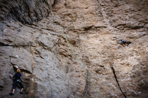 Two rock climbers scale a steep, vertical cliff face dotted with chalk marks and safety ropes in Bishop, California. One climber, wearing a black outfit, is near the bottom, while the other, in blue and black, is halfway up the rugged and towering rocky surface. visit bishop