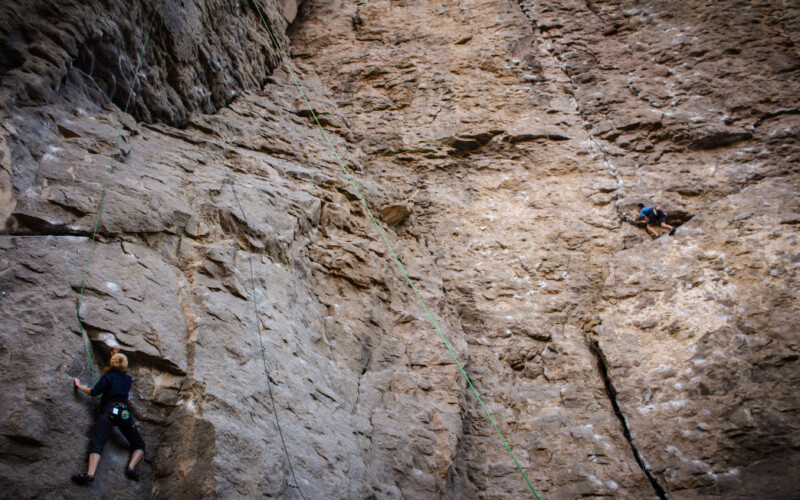 Two rock climbers scale a steep, vertical cliff face dotted with chalk marks and safety ropes in Bishop, California. One climber, wearing a black outfit, is near the bottom, while the other, in blue and black, is halfway up the rugged and towering rocky surface. visit bishop