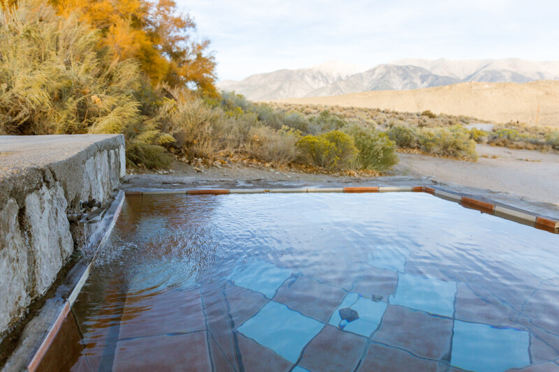 A serene hot spring pool with clear water, surrounded by rustic stone tiles, overlooks a rocky desert landscape with sparse vegetation in Bishop, California. In the distance, tall, rugged mountains rise under a bright, partly cloudy sky. visit bishop