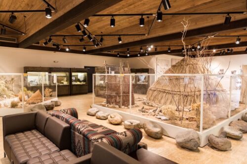 A museum exhibit features a traditional hut, nature dioramas, and seating with patterned blankets. visit bishop