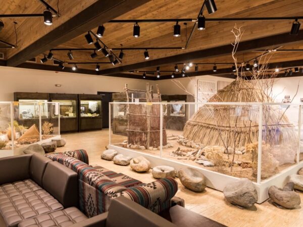 A museum exhibit features a traditional hut, nature dioramas, and seating with patterned blankets. visit bishop